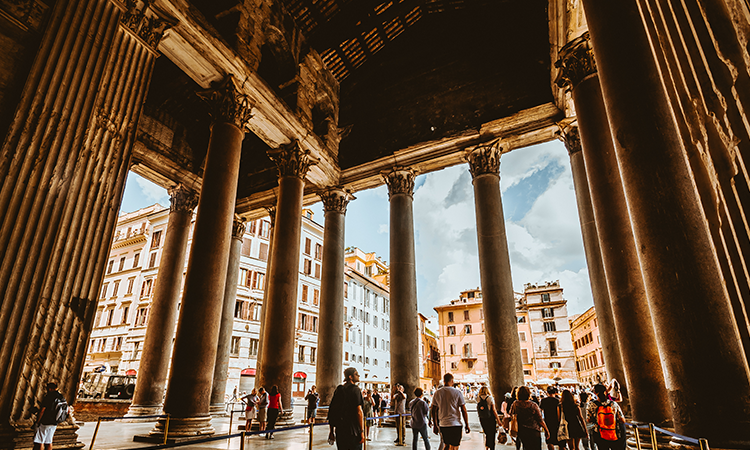 View from inside the Pantheon in Rome, looking out through its massive Corinthian columns toward the bustling square outside with visitors and historic buildings.