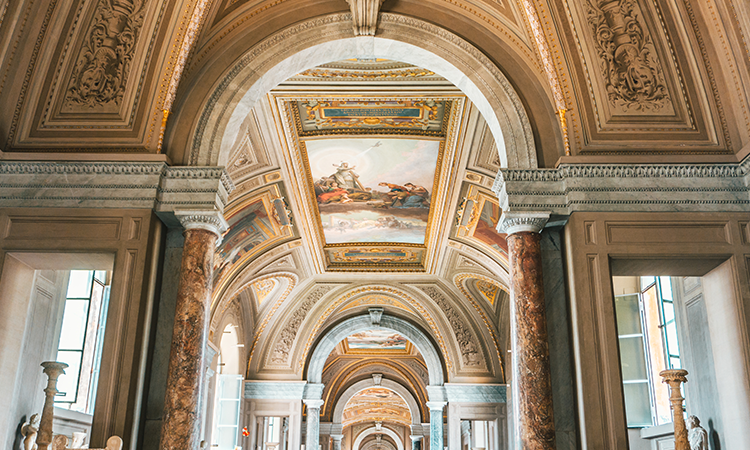 Ornate hallway inside the Vatican Museums, with marble columns, arched ceilings, and frescoed artwork.