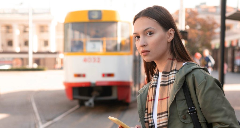 Young woman holding a phone while waiting at a tram stop, with a yellow and white tram approaching in the background.