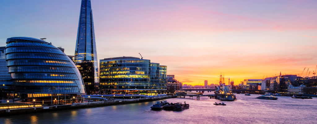 Panoramic view of London at sunset featuring the Shard, Tower Bridge, and modern riverside buildings along the Thames.