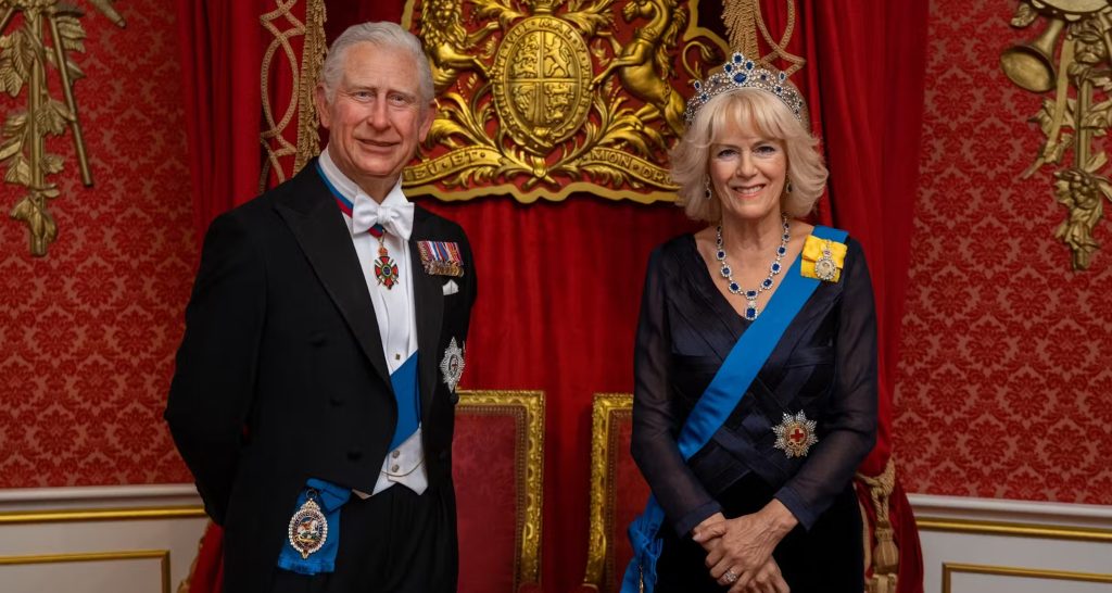 Wax figures of a formally dressed man and woman wearing ceremonial attire with sashes and medals, displayed in front of a red and gold backdrop at a museum.