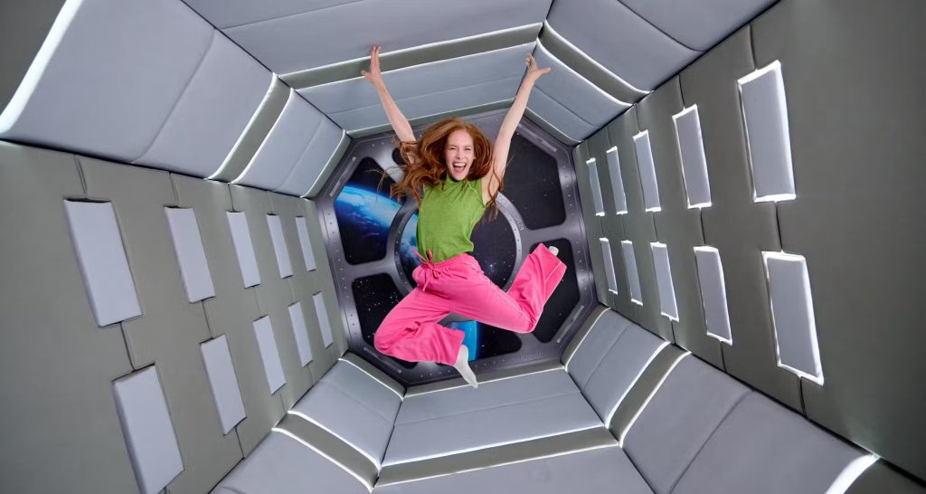Smiling girl jumping inside a futuristic, space-themed room with padded walls and a view of Earth through a window.