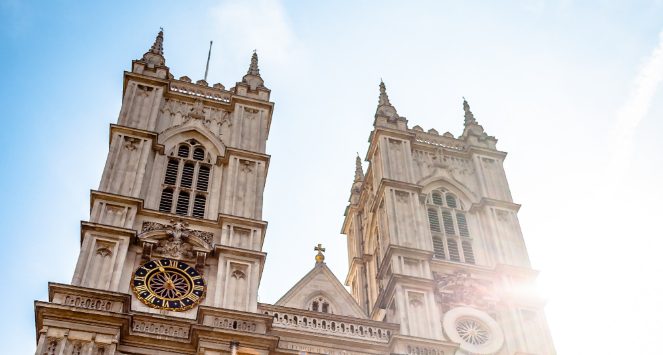 Close-up view of Westminster Abbey’s Gothic towers and clock face in London, with sunlight shining behind the historic church.